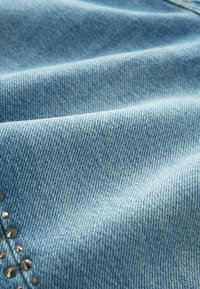 Țesătură din denim de un albastru deschis, cu o textură netedă, având o cusătură subtilă și mici accente metalice pe o margine.