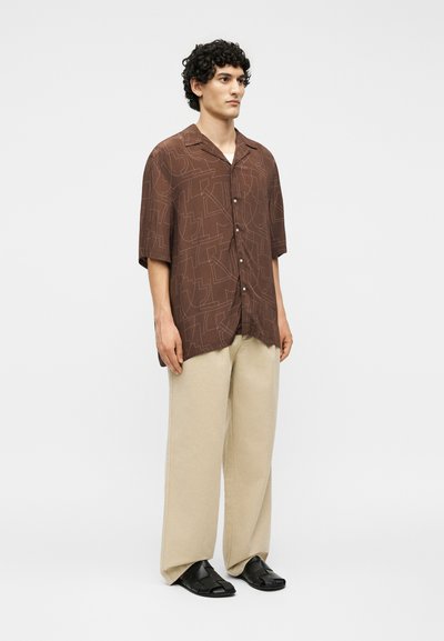 Άνδρας με καφέ πουκάμισο με σχέδια και κοντά μανίκια, μπεζ παντελόνι φαρδύ και μαύρα πλεκτά δερμάτινα παπούτσια, στέκεται μπροστά σε λευκό φόντο.