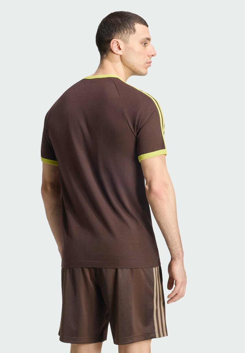 Mann mit dem Gesicht nach hinten, trägt ein braunes Sportshirt mit gelbem Besatz und passende braune Shorts mit beigen Streifen.