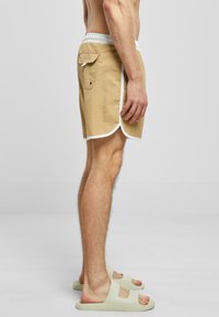 Beige svømmebukser med hvit kant, elastisk liv, og baklomme. Båret med lyse sandaler. Sidevisning som viser knelange design.