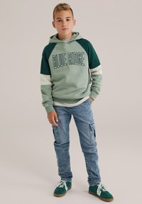 Zeleno-siva kapuca s napisom "BLUE RIDGE", u kombinaciji s plavim cargo trapericama i zelenim tenisicama, u ležernom stilu.