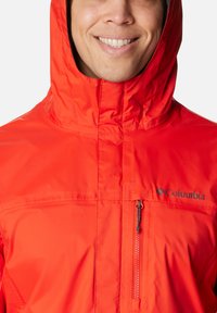 Leuchtend orangefarbene wasserdichte Jacke mit Kapuze, ausgestattet mit einem Frontreißverschluss und einer kleinen Brusttasche. Logo auf der oberen linken Seite.