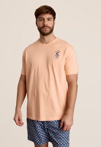 Perzik kleurige katoenen t-shirt met korte mouwen en een kleine ijsje-illustratie op de linkerkant van de borst. Ggedragen met blauwe geprinte shorts.