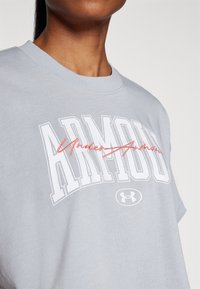 Harmaa puuvillapaita, jossa on valkoinen "ARMOUR" -logo ja punainen kursiivilla kirjoitettu "Under" -teksti yllään. Paita on lyhythihainen ja siinä on pyöreä kaula-aukko.