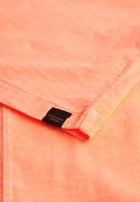 Tejido de camiseta de algodón color naranja coral con una superficie ligeramente texturizada. Incluye una etiqueta de marca negra cerca del dobladillo.