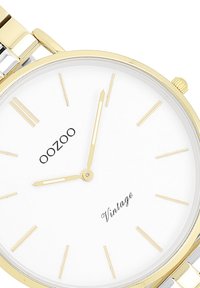 Montre dorée avec un cadran blanc, présentant des marqueurs d'heure minimalistes, un bracelet argenté, et "OOZOO Vintage" imprimé en dessous de l'heure 12.