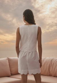 Haut sans manches blanc avec un ourlet festonné, associé à un short blanc. Le tissu semble léger et doux devant un arrière-plan de coucher de soleil flou.