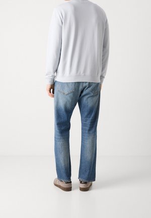 Mann trägt hellgraues Sweatshirt, blaue ausgewaschene Jeans und braune Schuhe, steht mit dem Rücken zum Betrachter vor weißem Hintergrund.