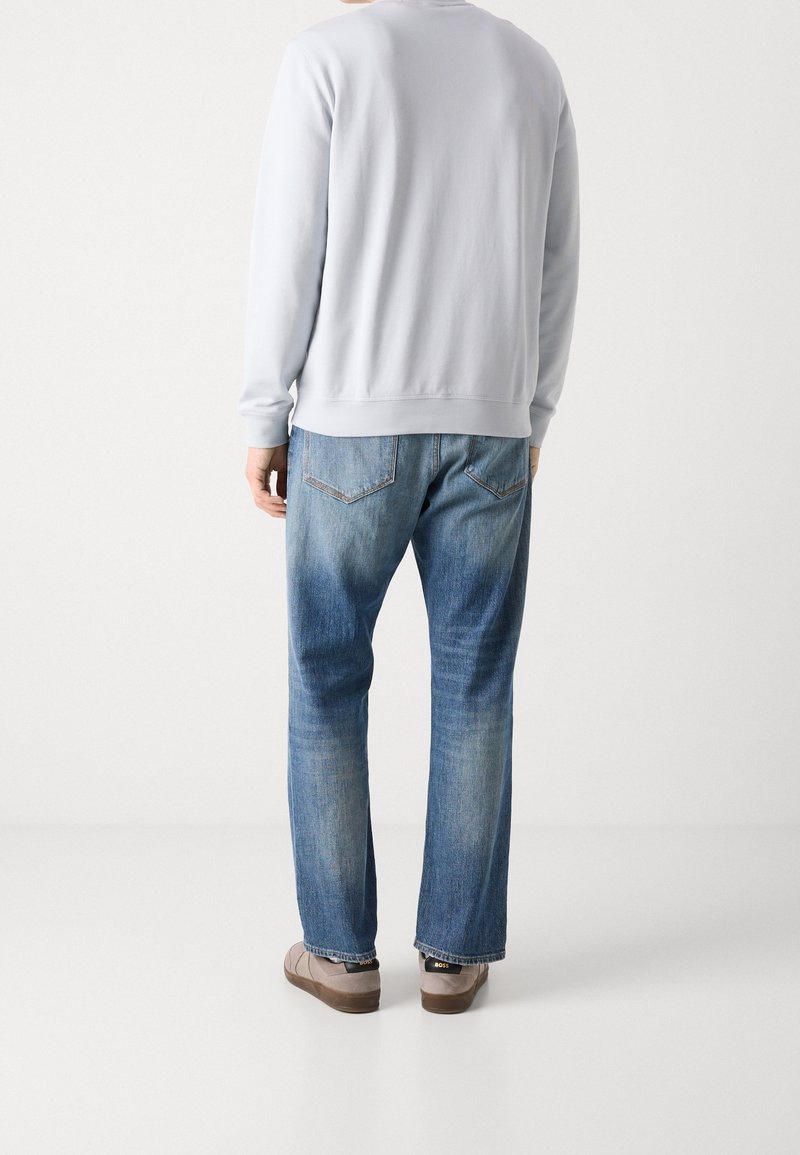 Homme portant un sweat-shirt gris clair, un jean bleu délavé et des chaussures marron, debout dos au spectateur devant un fond blanc.