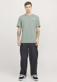 Grijs-groen T-shirt met ronde hals en korte mouwen, voorzien van een wit logo op de borst, gecombineerd met losse, donkere denim jeans en zwarte sneakers.