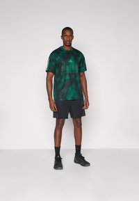 Green camouflage sports jersey with mesh texture, black shorts, black socks, and sneakers. Features three white stripes on the sleeves.