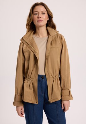 Kobieta z długimi włosami, ubraną w beżową bluzę rozpinaną na suwak nałożoną na beżową koszulę oraz niebieskie spodnie, stojąca na tle jasnego, jednolitego tła.