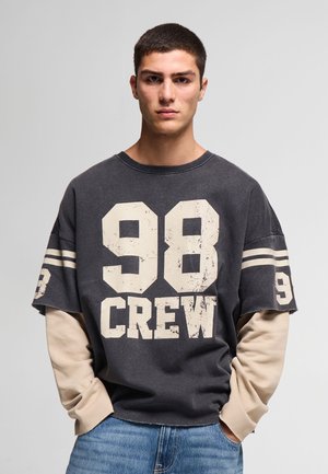 Giovane uomo con capelli corti che indossa una camicia nera oversize con testo "98 CREW" sovrapposto su maniche lunghe beige e jeans blu.
