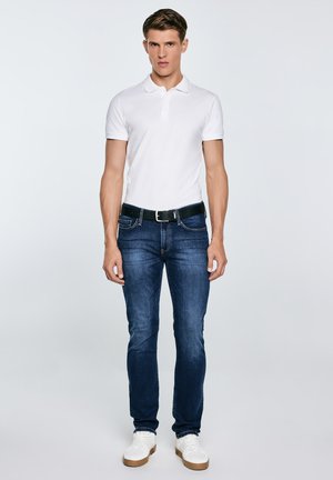 Mees seisab, kandes valget lühikeste varrukatega polosärki, tumelõhnavat teksapükse musta vöökinnitusega ja valgeid vabaajajalatsid, taustaks hele ühtlane taust.