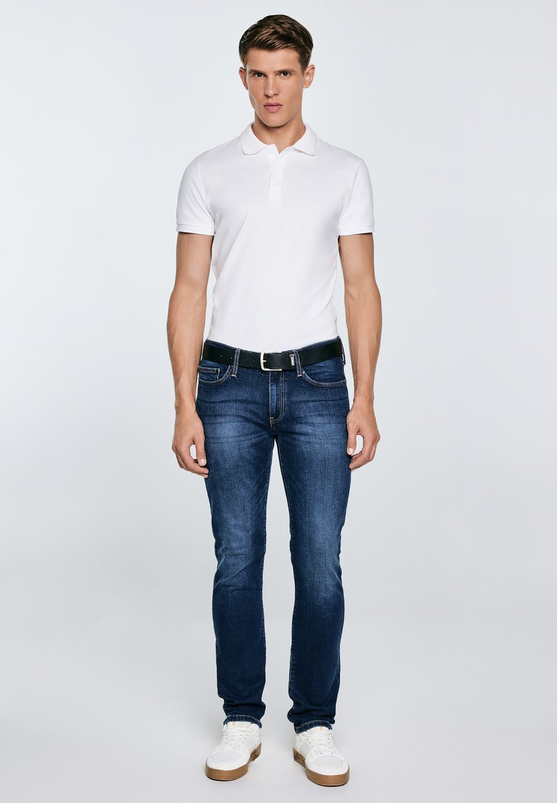 Mees seisab, kandes valget lühikeste varrukatega polosärki, tumelõhnavat teksapükse musta vöökinnitusega ja valgeid vabaajajalatsid, taustaks hele ühtlane taust.