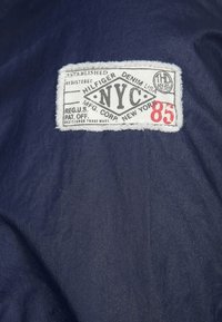 Tissu bleu marine avec une étiquette rectangulaire blanche portant "HILFIGER" et "NYC" en noir, "85" en rouge, ainsi que des détails supplémentaires de marque déposée.