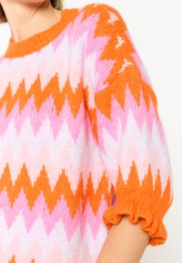 Megztinis su zigzago raštu rožinės, oranžinės ir baltos spalvų. Apykaklė su apvaliu iškirpimu ir banguotais oranžiniais rankogaliais. Minkšta, pūkuota tekstūra.