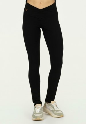 Vrouw die zwarte leggings met hoge taille en lichtgrijze sneakers draagt, staand tegen een effen achtergrond.