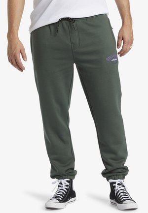 Pantalons de survêtement en coton vert avec une taille élastique et un cordon de serrage ajustable. Présente un logo violet sur la cuisse gauche et des chevilles resserrées.