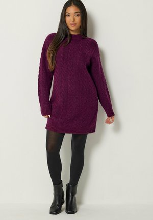Femme portant une longue robe-pull violette en maille torsadée, des collants noirs opaques et des bottines noires, debout devant un fond clair uni.