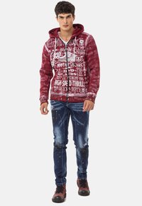 Rote Kapuzenjacke mit weißen grafischen Drucken, Reißverschluss vorne, Kängurutasche. Blaue Jeans mit Farbeffekt-Muster, getragen mit schwarz-roten Stiefeln.