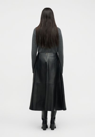 Γυναίκα με μακριά σκούρα μαλλιά, που φοράει γκρι μακρυμάνικη μπλούζα, μαύρη δερμάτινη midi φούστα και μαύρες μπότες με τακούνι, στέκεται με την πλάτη προς τα εμπρός.