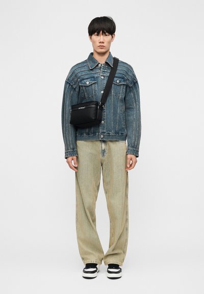 Νέος άνδρας που φοράει μπλε τζιν μπουφάν, άνετα ανοιχτόχρωμα τζιν, μαύρη τσάντα χιαστί και ασπρόμαυρα αθλητικά παπούτσια, στέκεται μπροστά σε απλό φόντο.