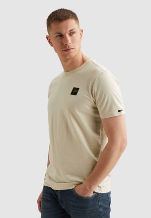 Jonge man die een beige T-shirt met korte mouwen draagt met een zwart logo, poseert met één hand in zijn zak en kijkt naar links.