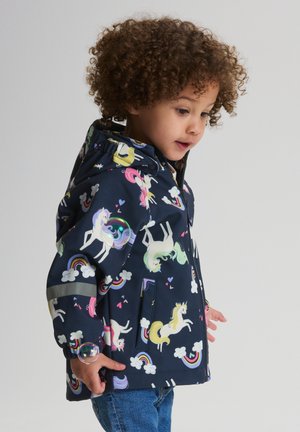 Curly-haired child wearing a navy raincoat with colorful unicorn, rainbow, and cloud patterns, holding a large soap bubble near their hand.