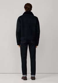 Donkerblauwe suedjacket met een groot bontkraag, gedragen met donkere spijkerbroeken en bruine schoenen. De jas heeft een rechte snit.