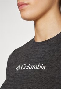 Tummanharmaa lyhythihainen paita, jossa on pyöreä kaula-aukko. Paidan rintamuksessa on valkoinen "Columbia"-logo, ja se on valmistettu teksturoidusta kankaasta.