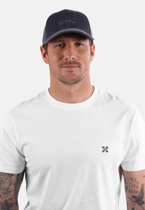 T-shirt en coton blanc à manches courtes et col rond, avec un petit design noir sur la poitrine gauche. Casquette bleu denim avec broderie.