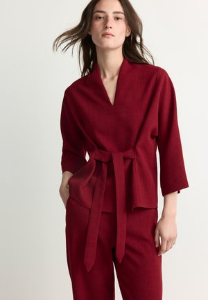 Young woman wearing a deep red wrap jacket with matching pants, standing with one hand in pocket against a plain background.