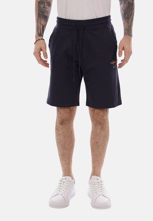 Hombre con pantalones cortos negros con cordón, zapatillas blancas y tatuajes visibles en el antebrazo, de pie con los brazos relajados a los lados.