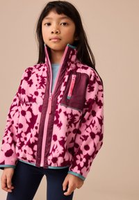 Gyapjúdzseki rózsaszín és lila virágmintákkal, magas gallérral, teljes cipzárral és egy mellzsebbel, amelyen rózsaszín cipzárhúzó található.