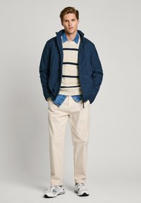 Marinblå pufferjacka över en beige randig tröja, komplett med en denimskjorta och krämfärgade byxor. Svart och vita sportskor.