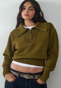 Frau mit langen, dunklen Haaren, die einen olivgrünen Zip-Hoodie, ein weißes T-Shirt darunter, dunkle Jeans und einen genoppten Gürtel trägt, posiert mit den Händen in den Taschen.