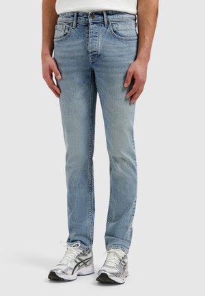 Man draagt lichtblauwe slim-fit jeans en witte sneakers, staand tegen een effen lichte achtergrond.