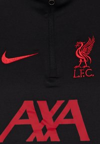 Crni sportski top s ovratnikom na zip, s crvenim Nike swooshom, logotipom Liverpool FC-a i velikim crvenim AXA natpisom sprijeda.
