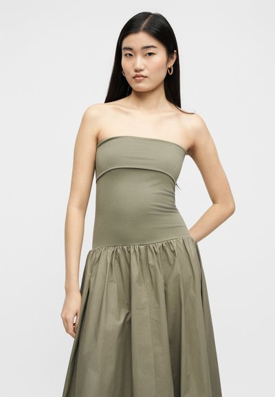 Νεαρή γυναίκα με μακριά σκούρα μαλλιά, που φοράει σκουλαρίκια κρίκους και ένα αμάνικο φόρεμα ελιάς με σφηκολεκτικό πέπλο, στέκεται μπροστά σε απλό φόντο.