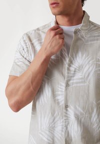Camisa de lino de manga corta en color beige claro con patrones de hojas blancas, que cuenta con un cierre de botones y un ajuste casual y relajado.