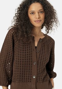 Cardigan en tricot brun présentant un motif de tissage lâche et ouvert, un col rond et trois grands boutons. Tissu texturé, léger avec une coupe décontractée.