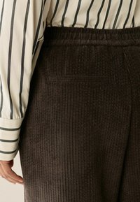 Braune Cordhosen mit einem strukturierten Rippmuster, ausgestattet mit einem elastischen Bund und einer einzelnen Gesäßtasche. Sichtbares weiß gestreiftes Hemd.