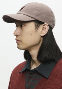 De pet heeft een zachte roze stof met een gebogen rand en geborduurde details. Het model draagt een donker overhemd met kraag onder een rode cardigan.