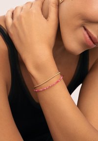 Pulseras de cuentas doradas y rosas llevadas en una muñeca. El tono de piel es bronceado. El dorado es elegante, mientras que las cuentas rosas son redondas y brillantes.