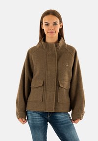 Giacca di lana marrone con colletto alto, dotata di due tasche frontali e dettagli di cucitura visibili, abbinata a jeans in denim blu.