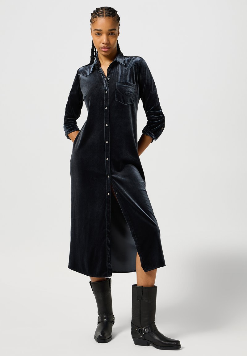 Abito camicia in velluto blu navy profondo con maniche lunghe, tasca sul petto, chiusura a bottoni e spacco laterale; abbinato a stivali neri in pelle al ginocchio.
