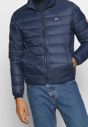 Man draagt een marineblauwe doorgestikte jas met een voorrits en zijzakken, gecombineerd met een blauwe spijkerbroek, hand in de jaszak.