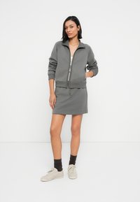 Γκρι μπουφάν με φερμουάρ, με ριμπ μανσέτες και τελείωμα, σετ με εφαρμοστό φόρεμα ίδιας απόχρωσης. Το μοντέλο φοράει ανοιχτού χρώματος κάλτσες και μπεζ αθλητικά παπούτσια.