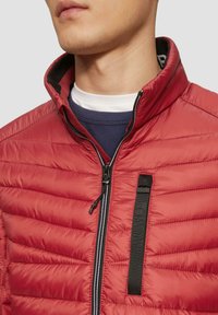 Giacca a vento rossa con cuciture orizzontali, colletto alto e chiusura con zip. Presenta una tasca verticale con una tracolla nera texturizzata.